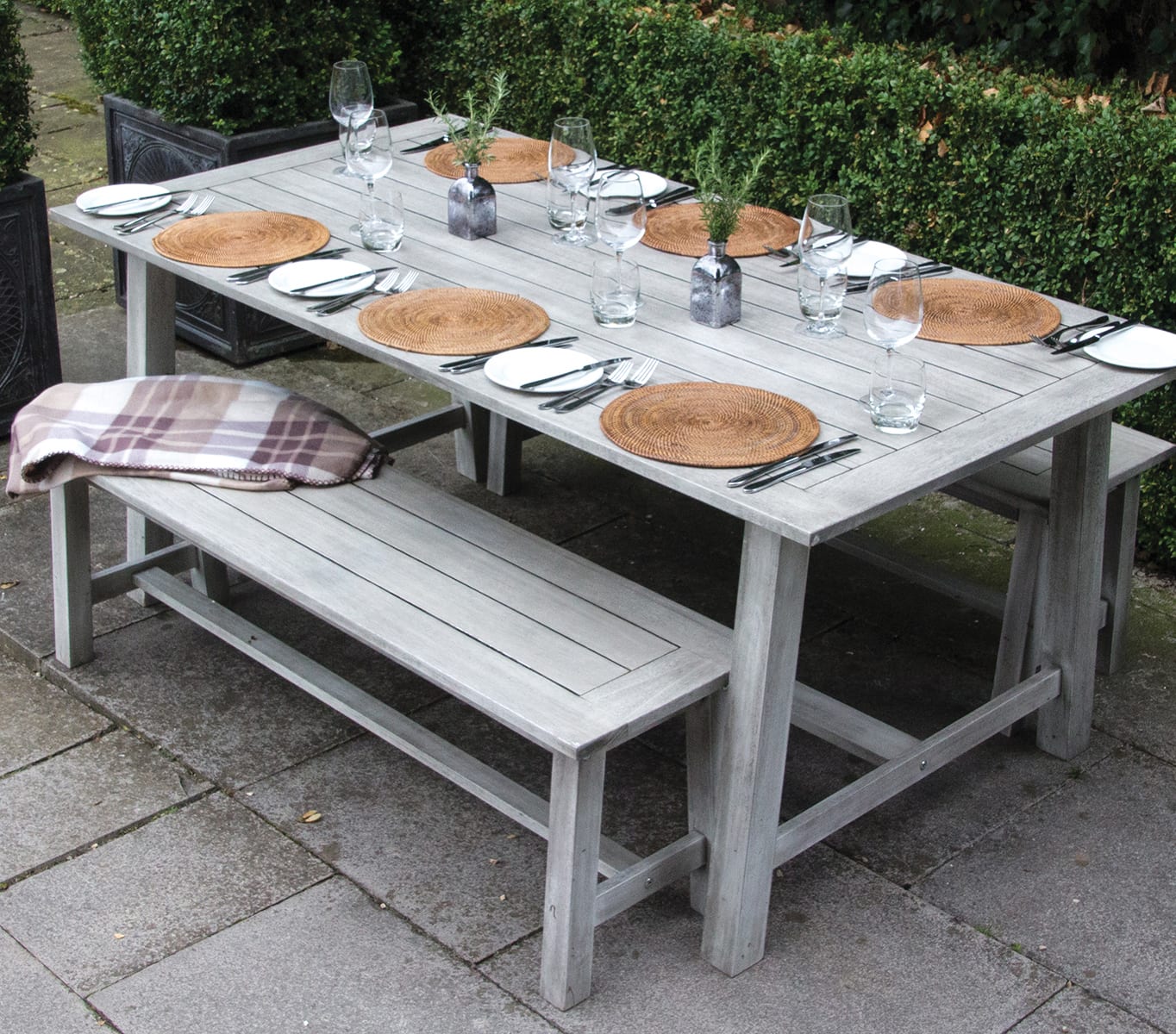 A wooden table and benches on a garden patio with six place settings.