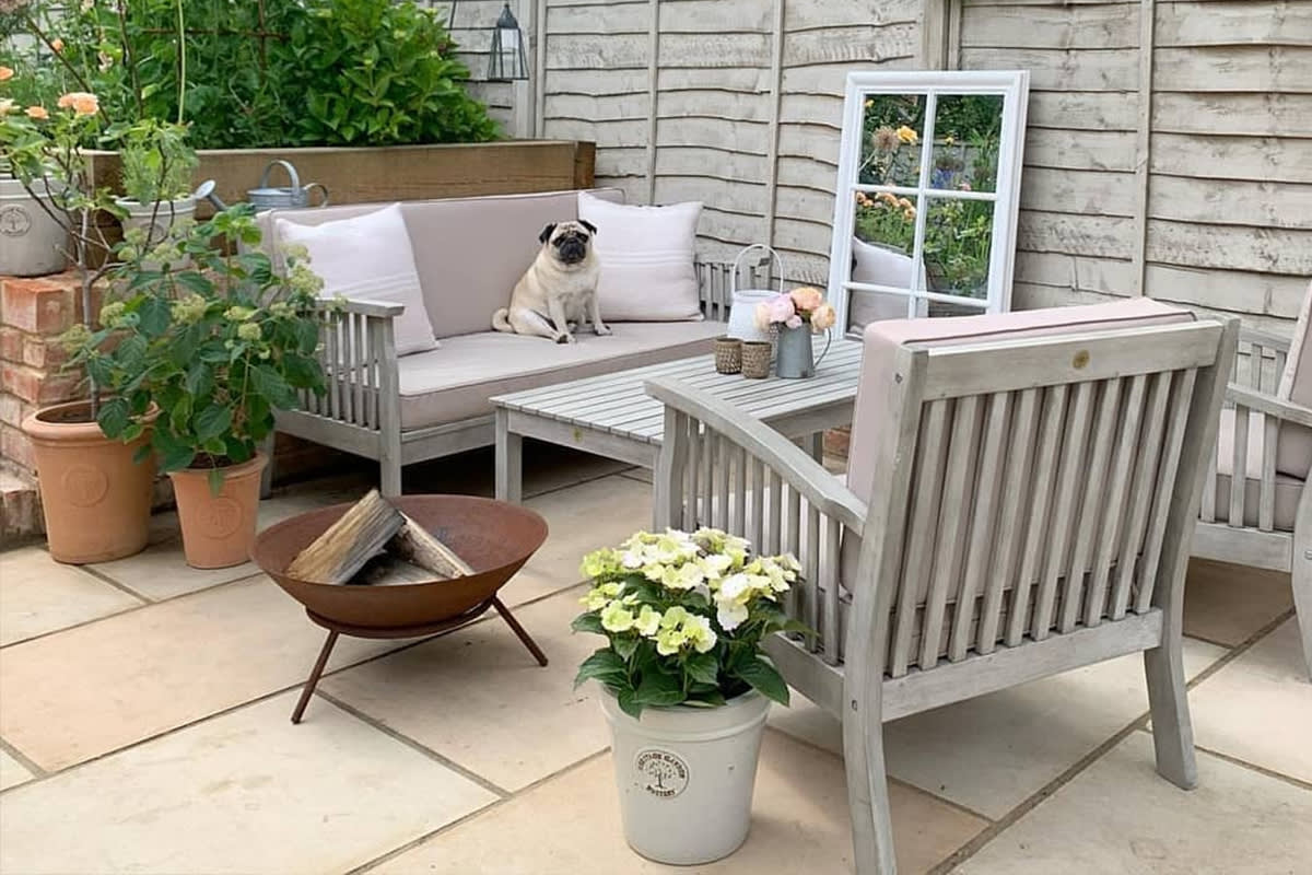 A Heritage Garden design wooden furniture set on a garden patio. Surrounded by flower pots, a fire pit, and a garden mirror. A pug sits comfortably on the sofa.